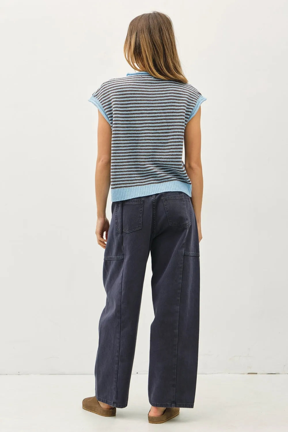 A woman wears a sleeveless knit mini striped piper button top with gray and light blue stripes paired with navy trousers