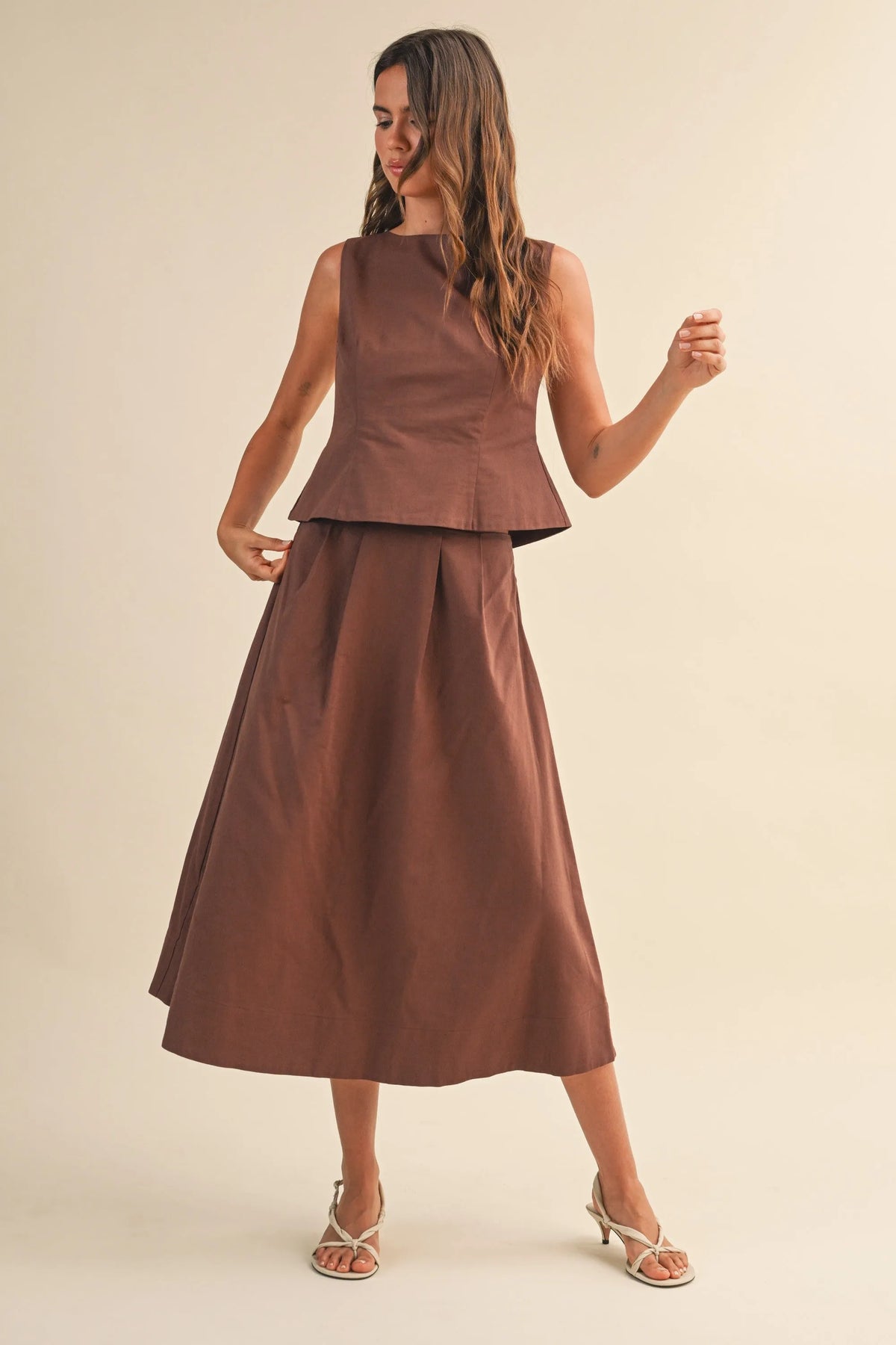 A woman wears a brown linen pleated waist flare midi skirt paired with a sleeveless peplum top