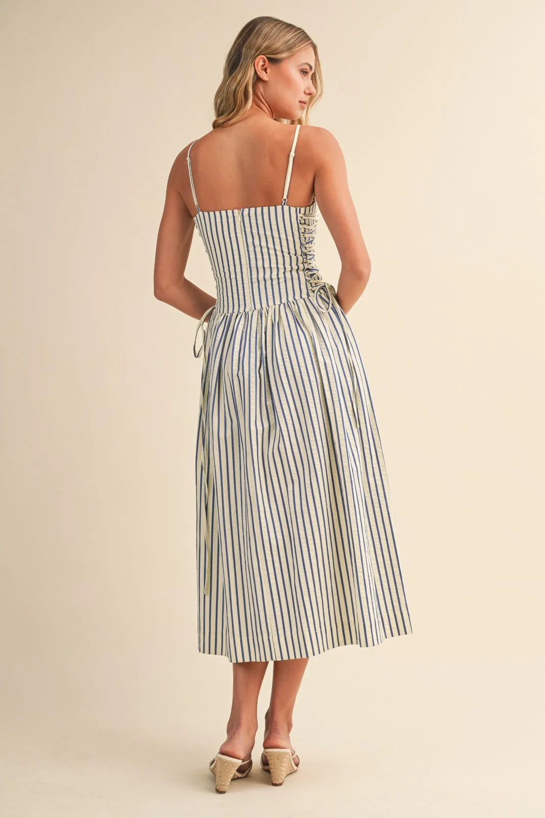 A woman wears a striped midi dress with thin straps and a side tie back lace-up detail