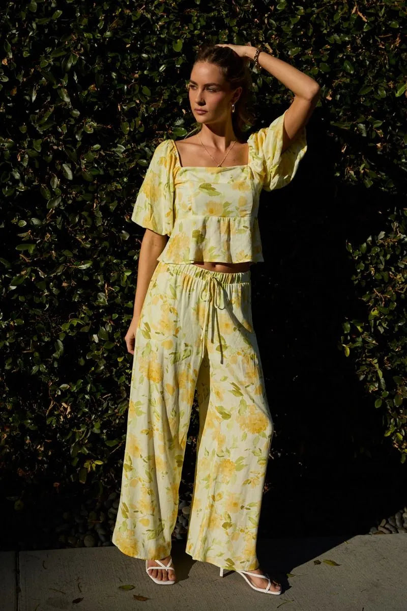 A woman wears soft yellow and green floral midi rise elastic waist band wide pants with lightweight fabric and ruffled sleeves