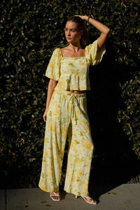 A woman wears soft yellow and green floral midi rise elastic waist band wide pants with lightweight fabric and ruffled sleeves