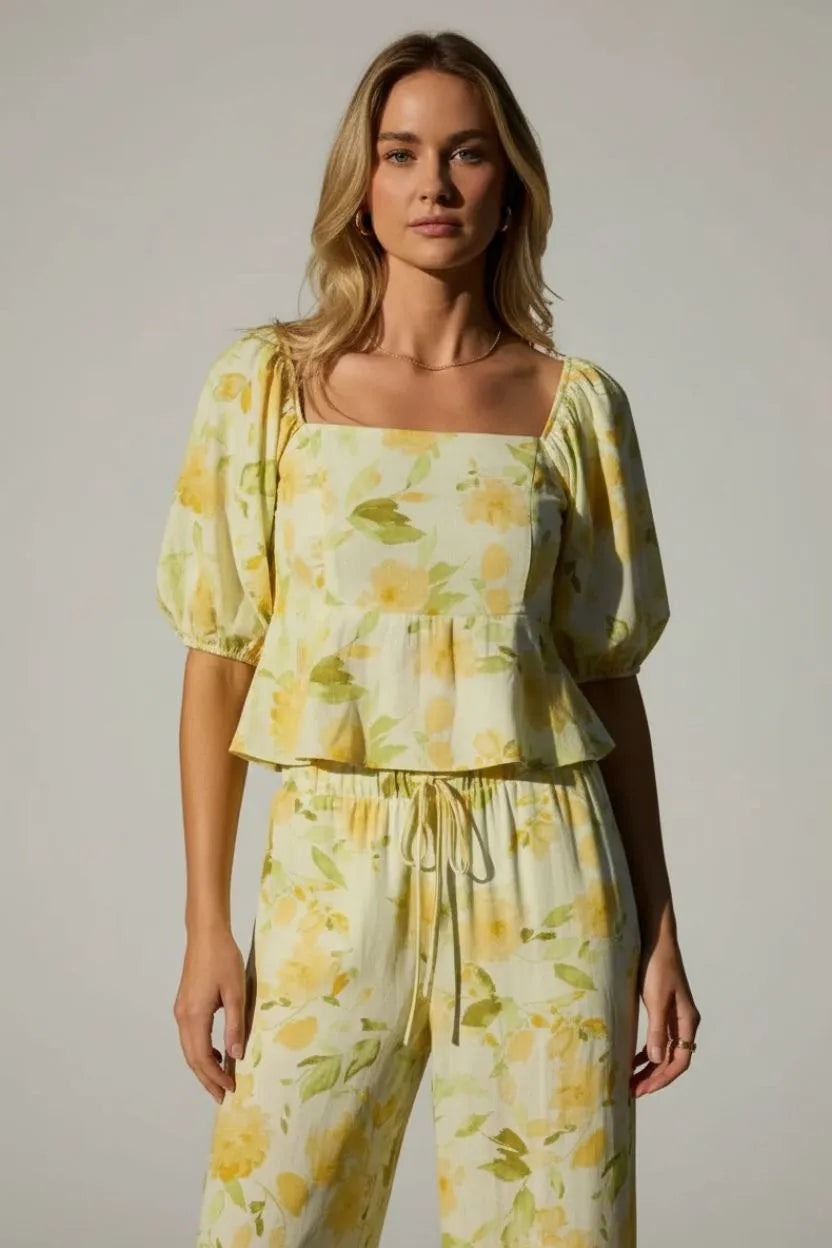 A woman wearing a vibrant yellow floral square neck bubble hem peplum blouse with matching drawstring pants on pale green
