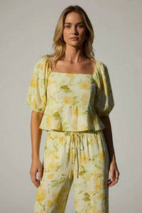A woman wearing a vibrant yellow floral square neck bubble hem peplum blouse with matching drawstring pants on pale green