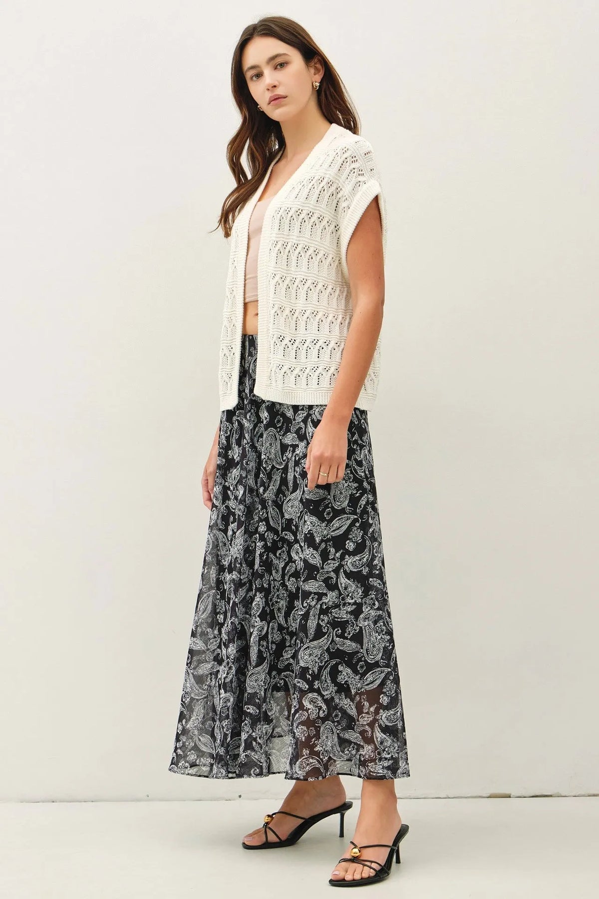 A woman wearing a white crochet knit boxy open cardigan with intricate lace patterns over a pink top and black skirt