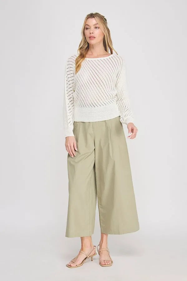 A woman wears a white hand yarn open stitch dolman sweater top with diagonal ribbing and olive wide-leg trousers
