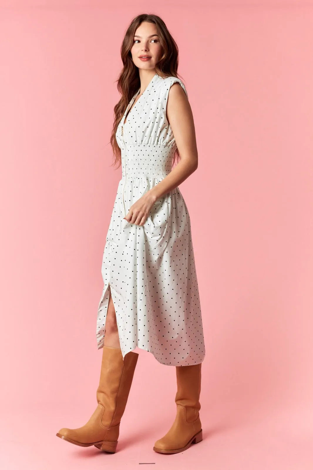 A woman wears a white pin polka dot flare midi dress with v-neck zip-up and fitted waist paired with tan boots