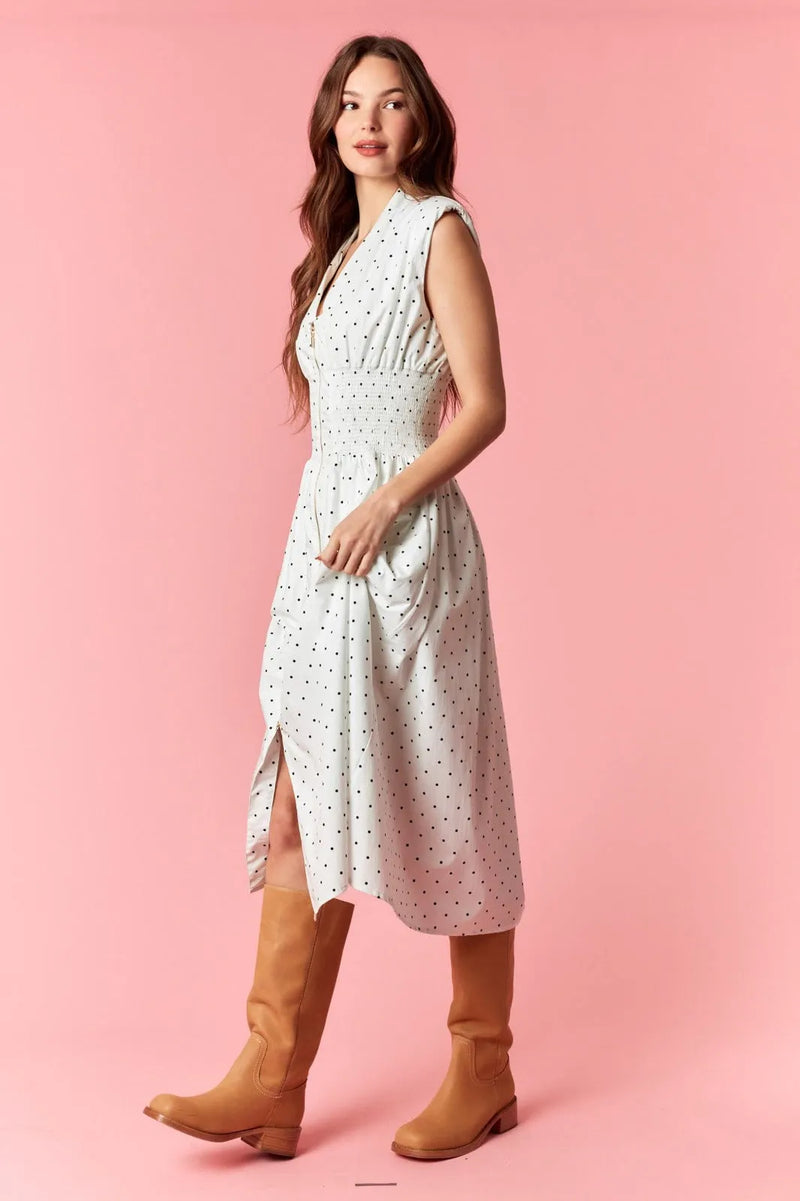 A woman wears a white pin polka dot flare midi dress with v-neck zip-up and fitted waist paired with tan boots