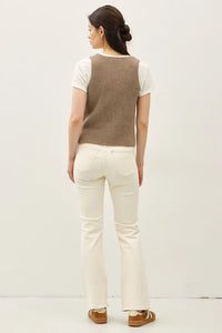 Light brown chunky V neck sweater vest with neck button over white top and off-white jeans