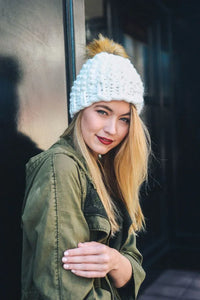 Person in white knit oversized chunky pom beanie and olive green jacket