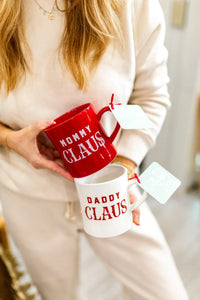 Two Christmas-themed Mommy Daddy Claus stoneware mugs held by a person in a cream sweater