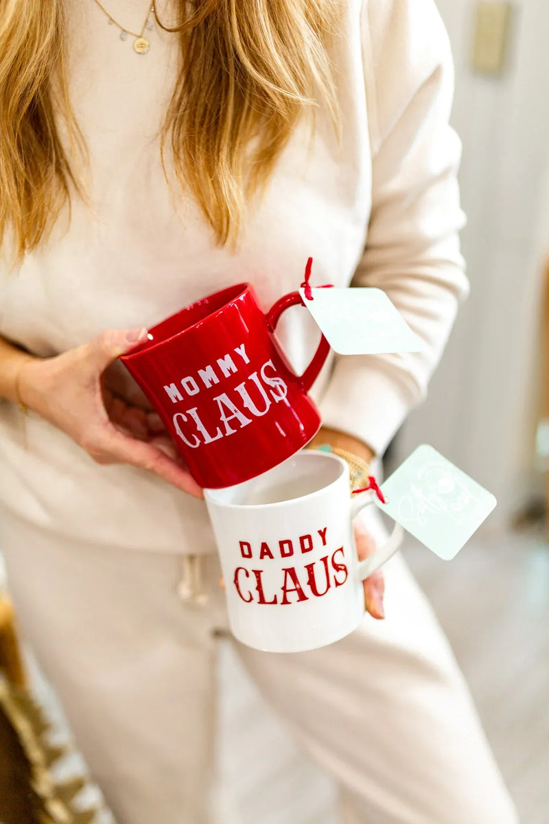 Two Christmas-themed Mommy Daddy Claus stoneware mugs held by a person in a cream sweater