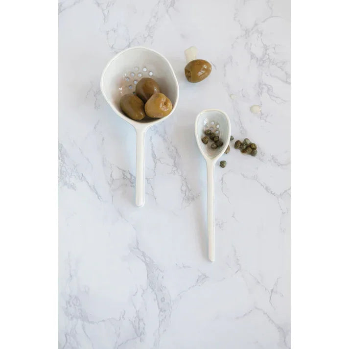 Two white bowls with olives placed on a marble counter beside a Stoneware Strainer Spoon