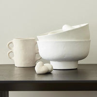 White ceramic bowls and cups on a surface with a Paper Mache Vase featuring handles