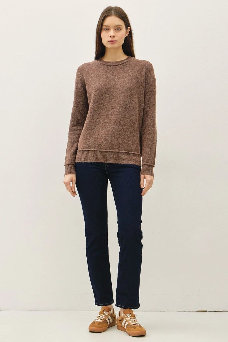 Woman modeling brown crew neck pullover with exposed seams, dark blue jeans, and tan sneakers