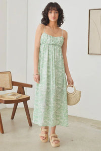 Woman in a green sleeveless shirring tropical print maxi dress standing by a chair