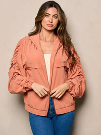 Woman in peach jacket with long ruched sleeves, zip pockets, and hooded design