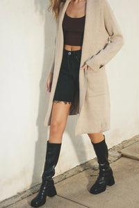 Woman wearing a cozy open long cardigan with pockets, black crop top, shorts, and knee-high boots