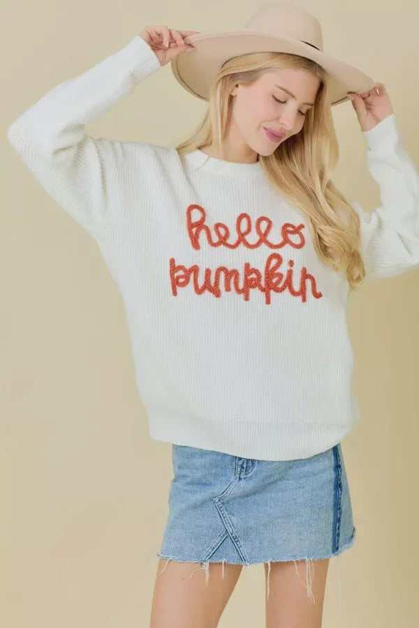 Woman in a white crew neck sweater with hello pumpkin text, denim skirt, and beige hat