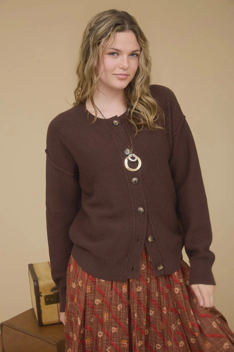 Woman wearing brown exposed seam button down knit cardigan with long necklace and patterned skirt