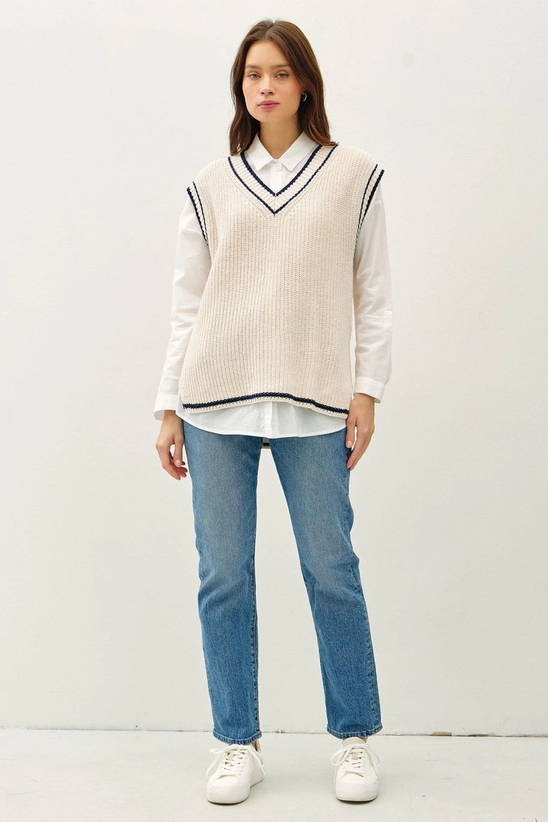 Cream-colored varsity style sweater vest with accent stripe layered over a white shirt and blue jeans