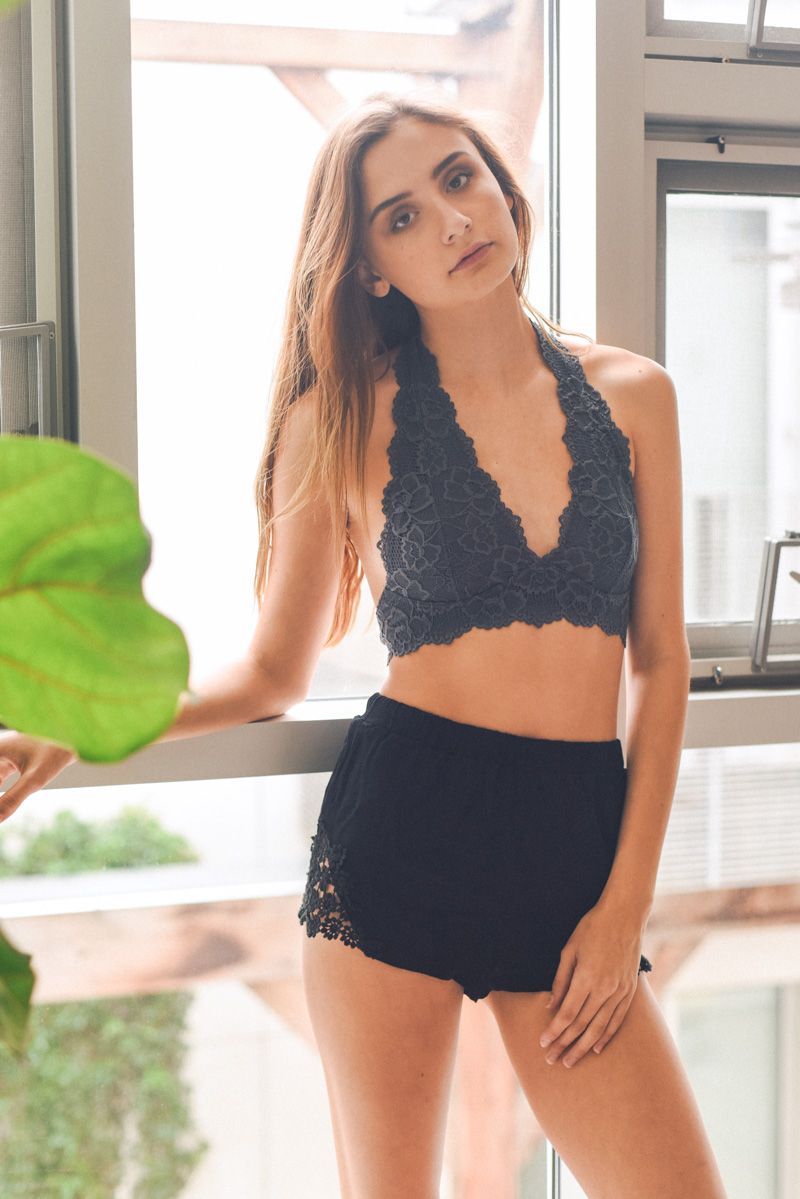 A girl standing against a window wearing a gray, lace, halter top bralette and black shorts.